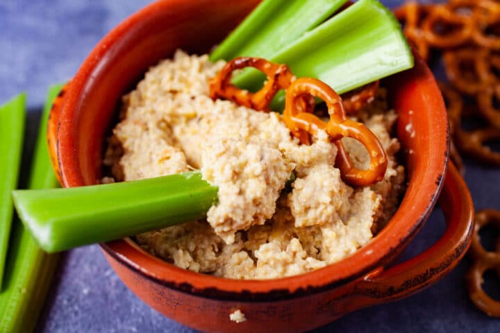  uma tigela de terracota cheia de homus de tremoço e coberta com pedaços de aipo e pretzels.