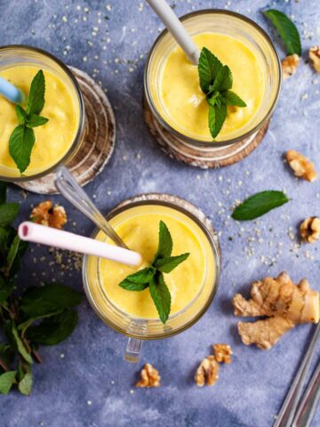 Three glasses filled with mango lassi, topped with mint leaves and each carrying a straw.