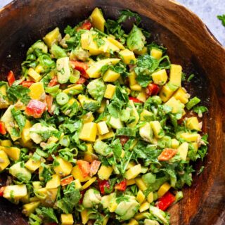 A large wooden bowl filled with fresh and creamy mango avocado salad, onions, cilantro, sunflower seeds and red bell peppers.