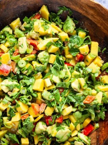 A large wooden bowl filled with fresh and creamy mango avocado salad, onions, cilantro, sunflower seeds and red bell peppers.