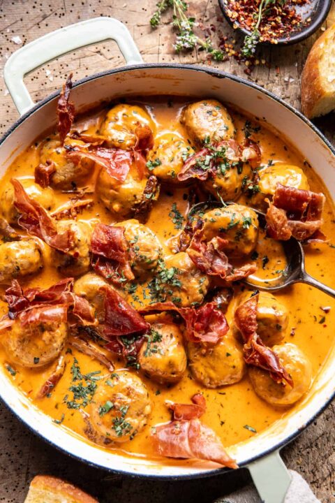 an overhead shot of meatballs in a white skillet in a creamy sauce with pieces of crispy prosciutto nestled around the meatballs.