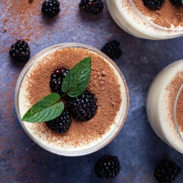 Glasses filled with mascarpone mousse, types with cacao powder and garnished with blackberries and mint leaves.