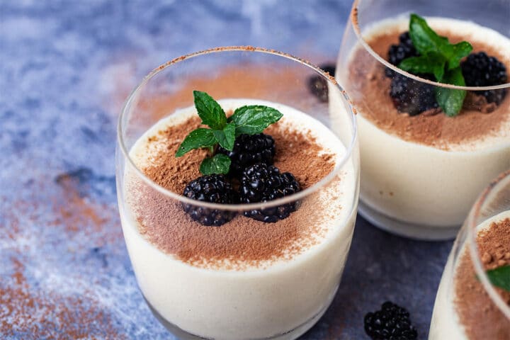 Glasses filled with mascarpone mousse, types with cacao powder and garnished with blackberries and mint leaves.