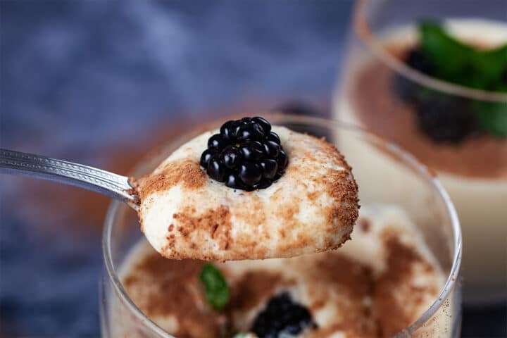 A small spoon holding up a light cacao coated mascarpone mousse, topped with a blackberry.