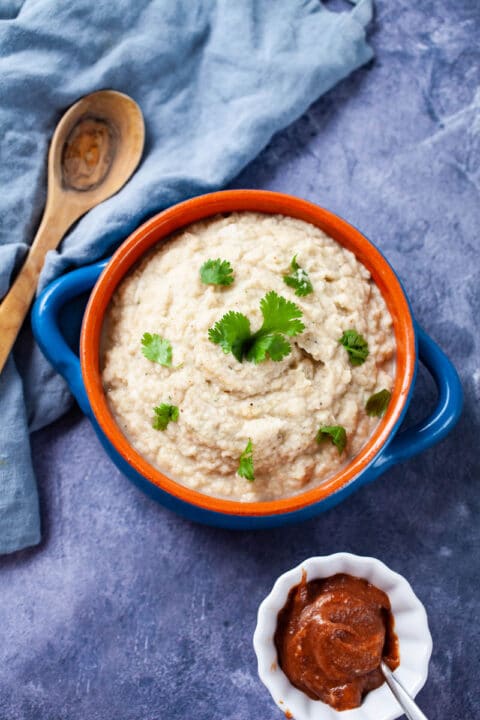 A terra cotta bowl filled with creamy mashed cauliflower and garnished with Parsley, next to a small bowl with apple butter.