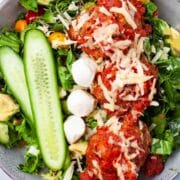 A meatball salad bowl with fresh cucumber slices, lettuce, greens, mini tomatoes, cheese and dressing.