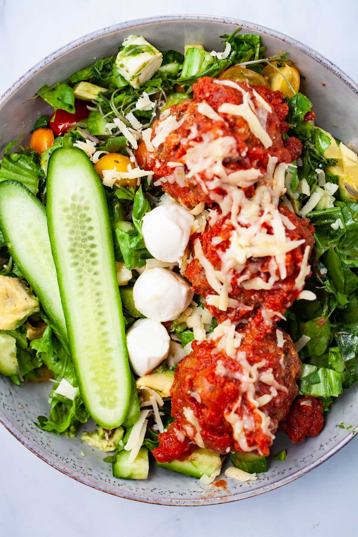 A meatball salad bowl with fresh cucumber slices, lettuce, greens, mini tomatoes, cheese and dressing.