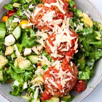 A meatball salad bowl with fresh cucumber slices, lettuce, greens, mini tomatoes, cheese and dressing.