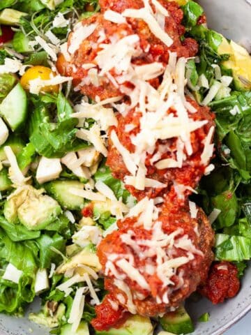 A meatball salad bowl with fresh cucumber slices, lettuce, greens, mini tomatoes, cheese and dressing.