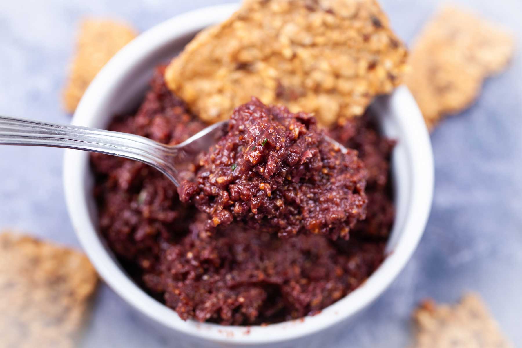 A spoon resting in a Kalamata olive and fig tapenade in a ramekin served with pieces of toast.