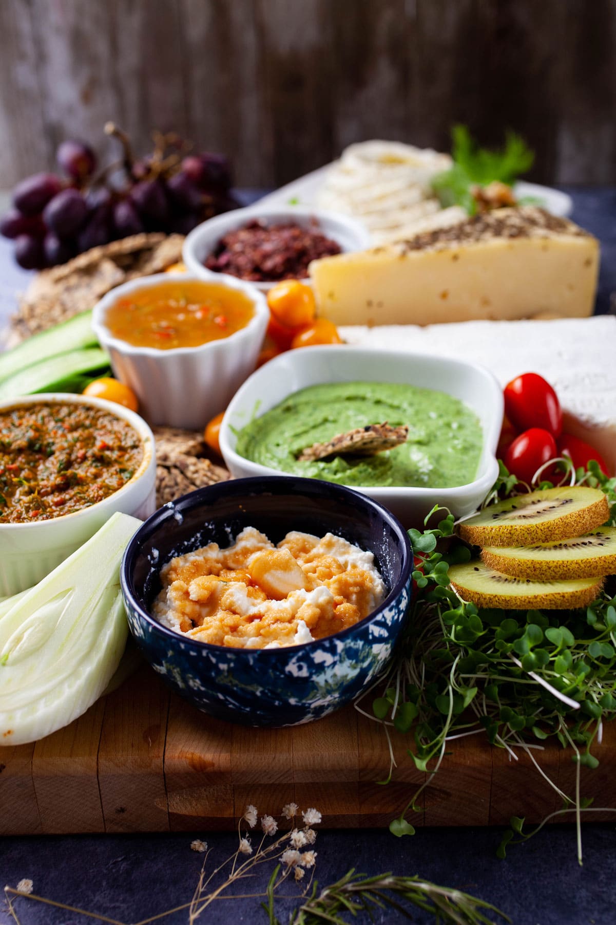 A wooden board topped with Italian dips, freshly cut veggies, fruits, cheeses, crackers, and spreads.