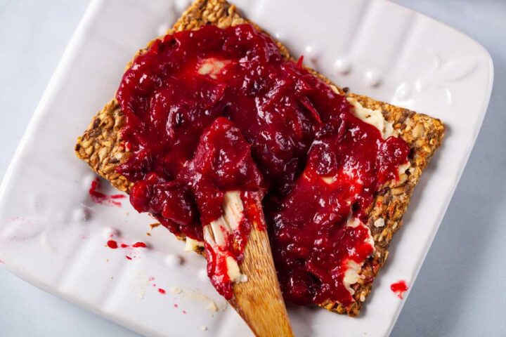 A wooden spoon smeared with butter and cranberry spread, resting on a smeared cracked seed cracker.