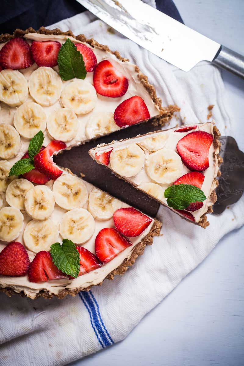 A slice cut and lifted from a round no-bake mascarpone cheesecake topped with sliced bananas and strawberries.