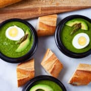 Three bowls of asparagus soup topped with avocado, half of a cooked egg and head of an asparagus with baguette bread on the side.