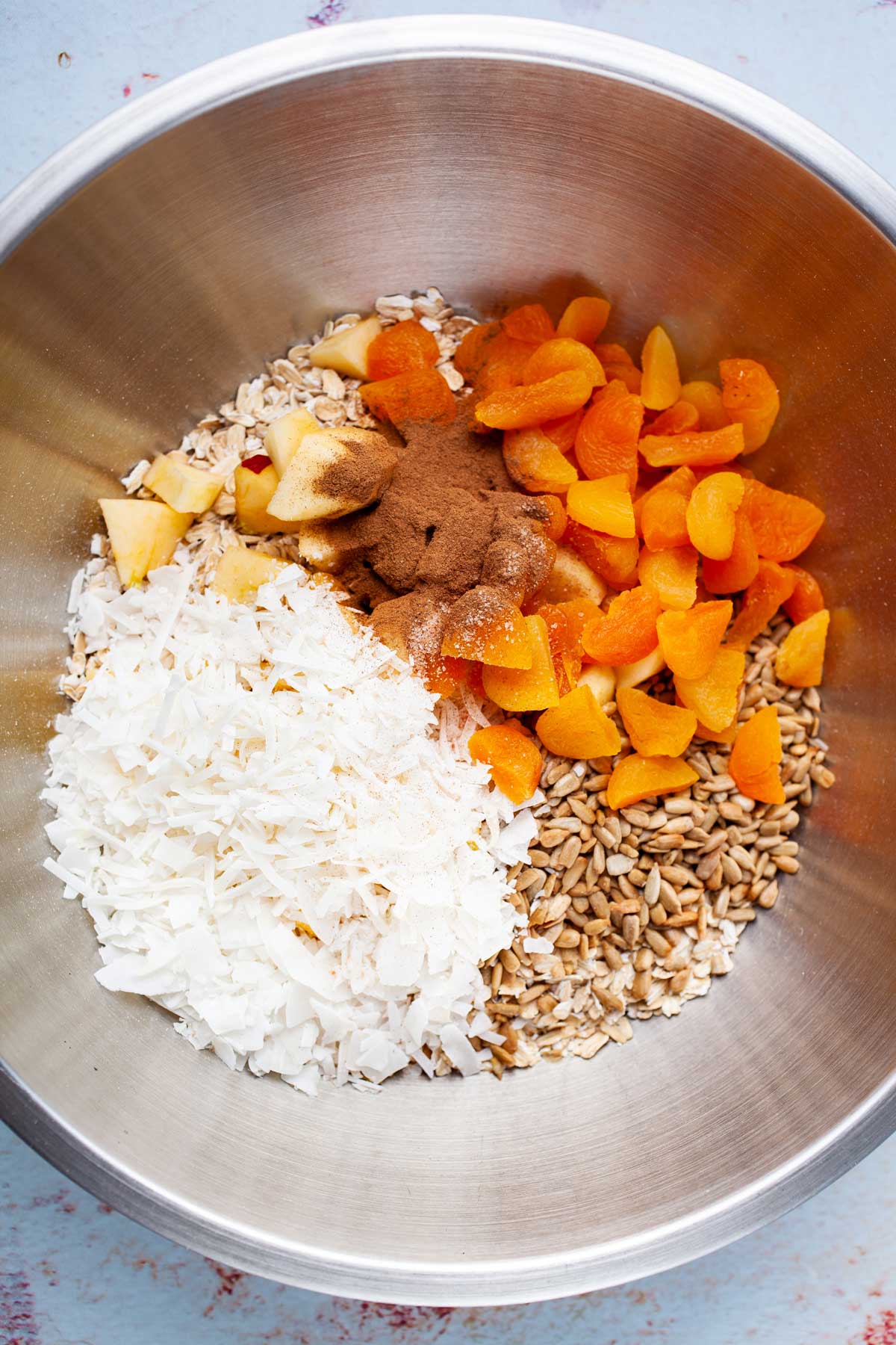 Oats, seeds, apples, dried fruits, coconut flakes, and spices in a large mixing bowl.