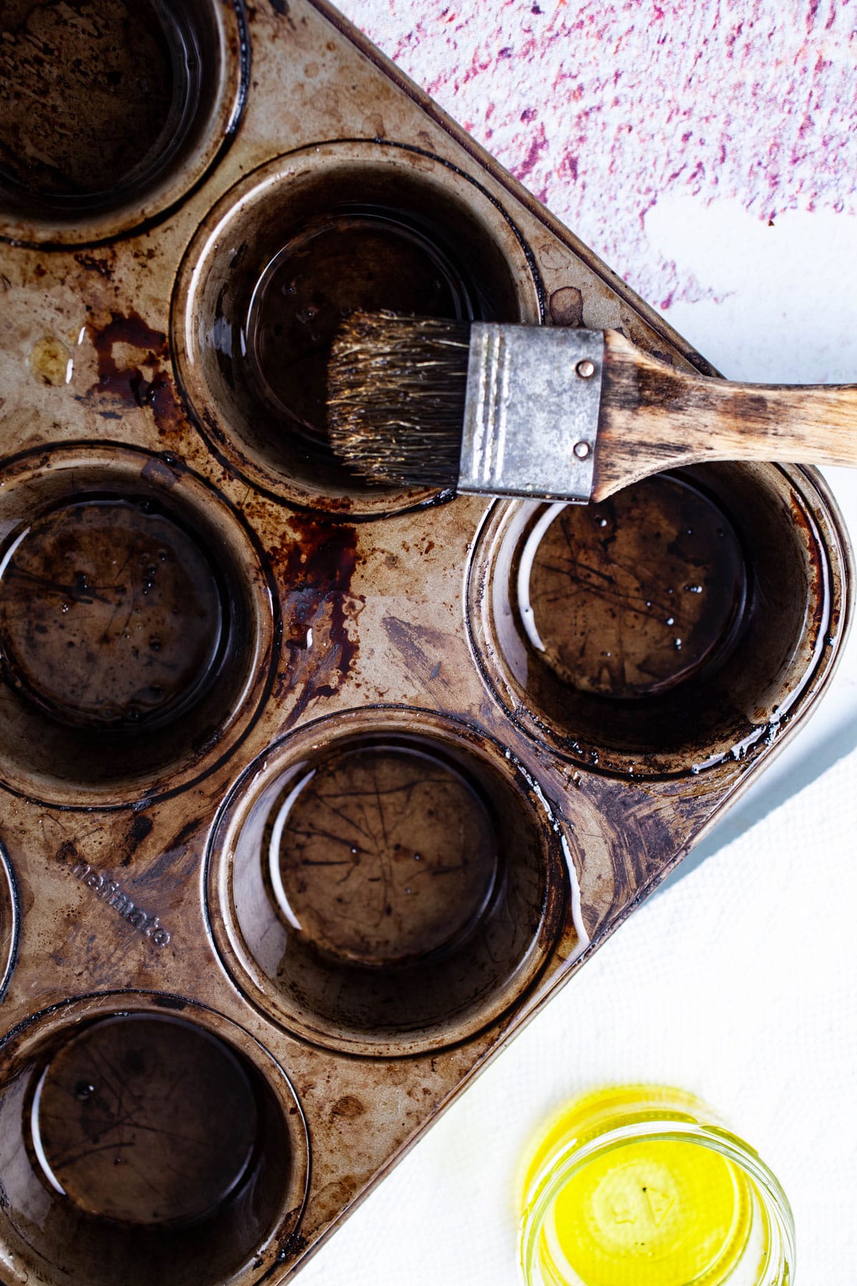A basting brush with oil resting on a tin muffin pan next to a small cup filled with olive oil.