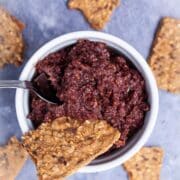 A spoon resting in a Kalamata olive and fig tapenade in a ramekin served with pieces of toast.