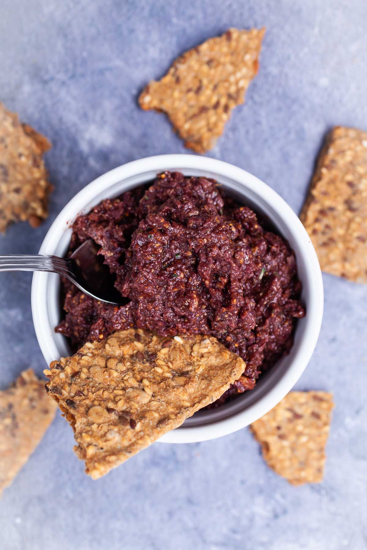 A spoon resting in a Kalamata olive and fig tapenade in a ramekin served with pieces of toast.