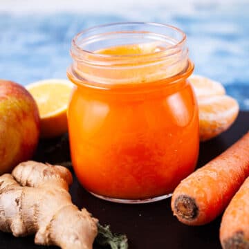 An orange carrot ginger juice in a glass served on a serving board with the ingredients in the background.