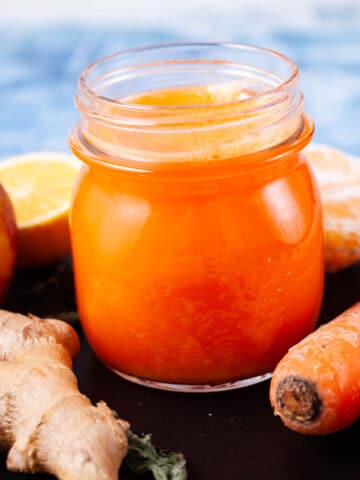 An orange carrot ginger juice in a glass served on a serving board with the ingredients in the background.