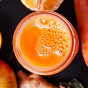 An orange carrot ginger juice in a glass served on a serving board with the ingredients in the background.