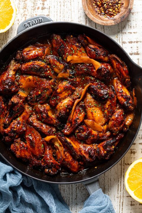 an overhead shot of spicy orange chicken wings in an enameled skillet with a blue napkin wrapped around the handle orange slices and crushed red pepper flakes are visible at the sides of the shot.