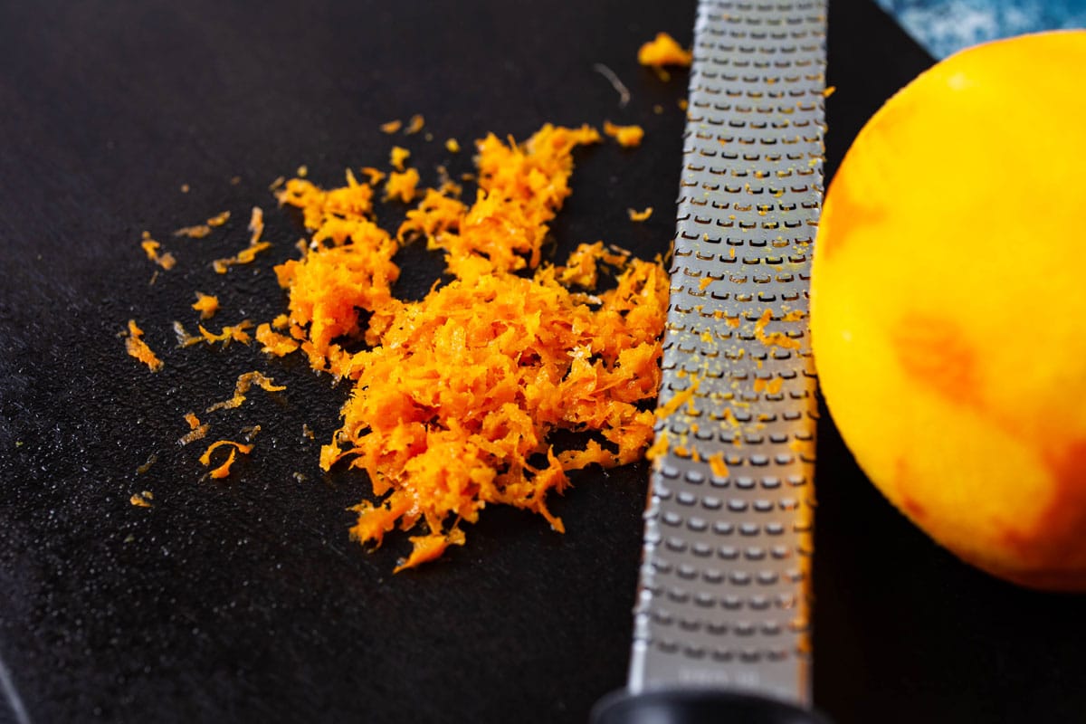 Orange zest and a zest grater next to a zested orange on a dark wooden board.