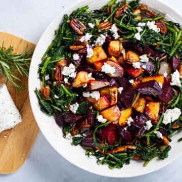 A large plate filled with roasted beets, potatoes, sauteed dandelion greens, pecans, next to a wooden plate topped with extra goat cheese and Rosemary.