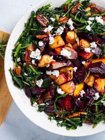 A large plate filled with roasted beets, potatoes, sauteed dandelion greens, pecans, next to a wooden plate topped with extra goat cheese and Rosemary.