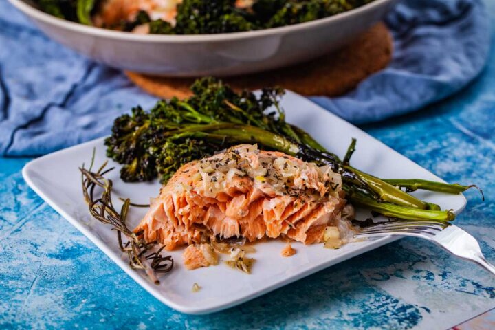 A piece of baked salmon served with baked baby broccoli on a plate, and garnished with thyme.