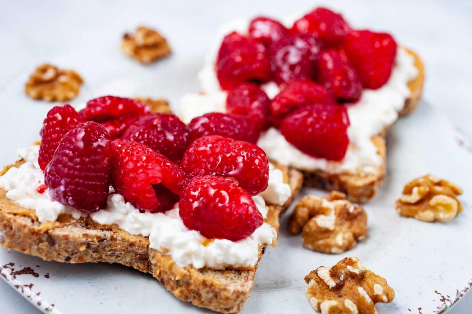 Two slices of whole-grain bread topped with cottage cheese and fresh raspberries are placed on a white plate, surrounded by scattered walnut pieces.