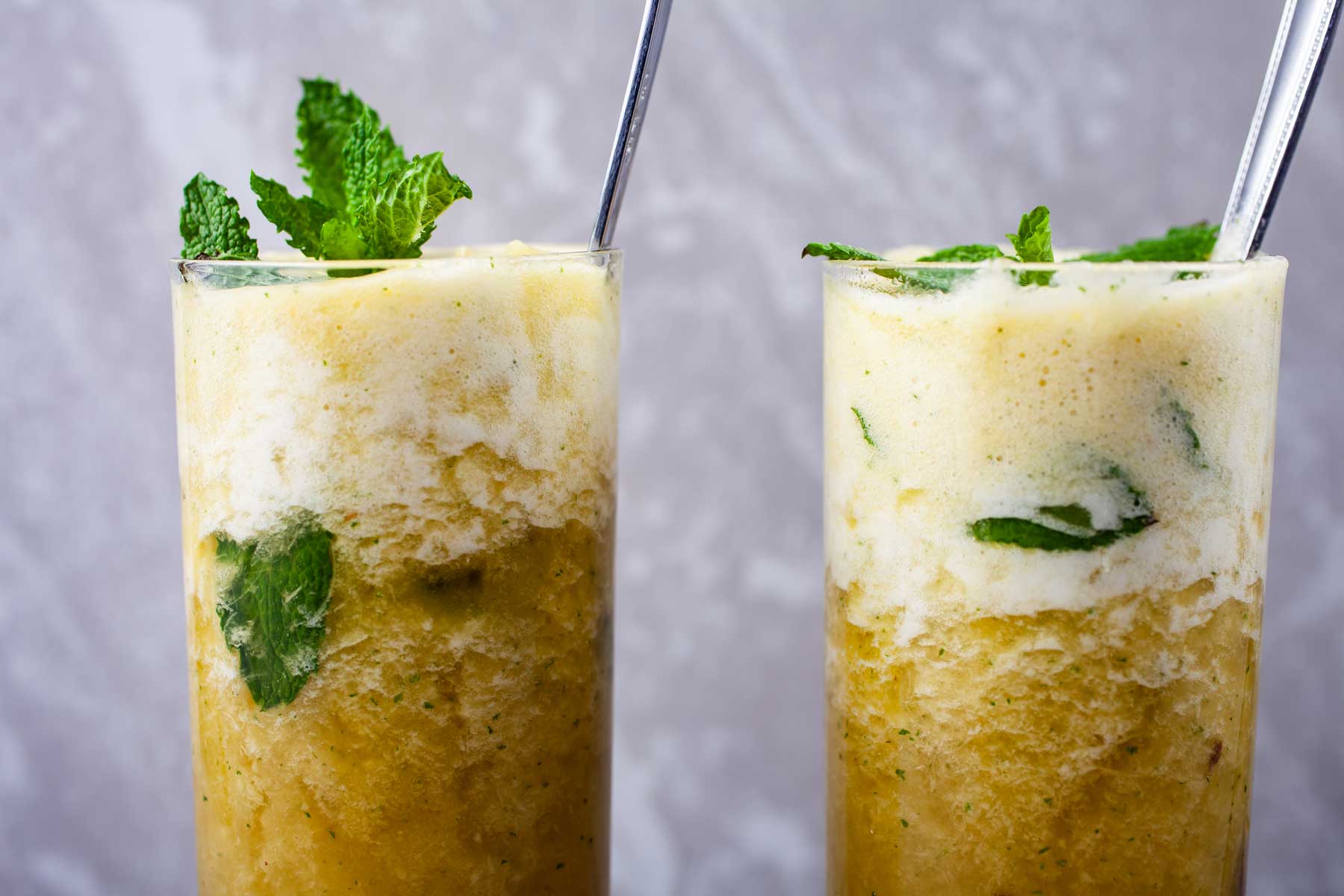 Large spoons in two sparkling pineapple mocktail drinks with mint garnishes in tall glasses.