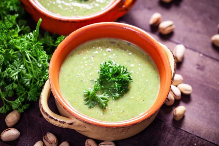 A close-up of a terra cotta bowl filled with green pea soup, topped with fresh Parsley leaves.