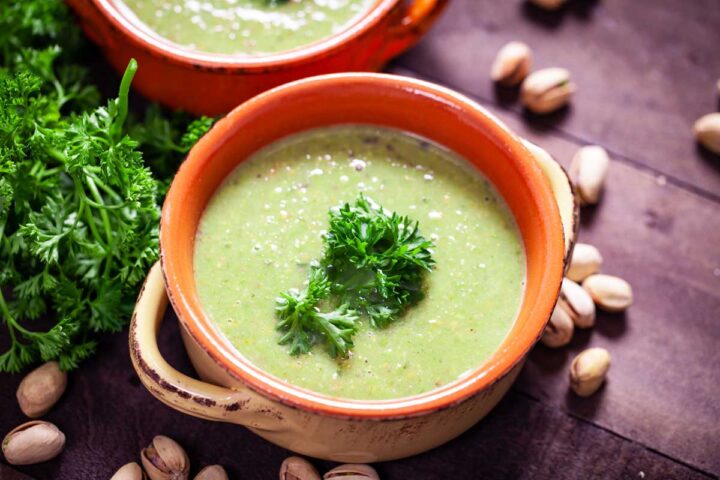 A close-up of a terra cotta bowl filled with green pea soup, topped with fresh Parsley leaves.