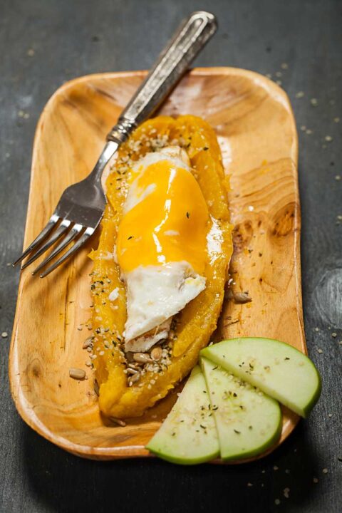 A large cooked and cross-wise opened plantain served on a wooden plate filled with a fried egg, nuts, and seeds and served with green apple slices on the side.