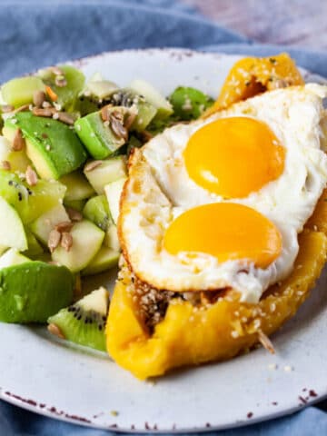 An opened cooked platano filled with eggs and seeds next to a green fruit salad.