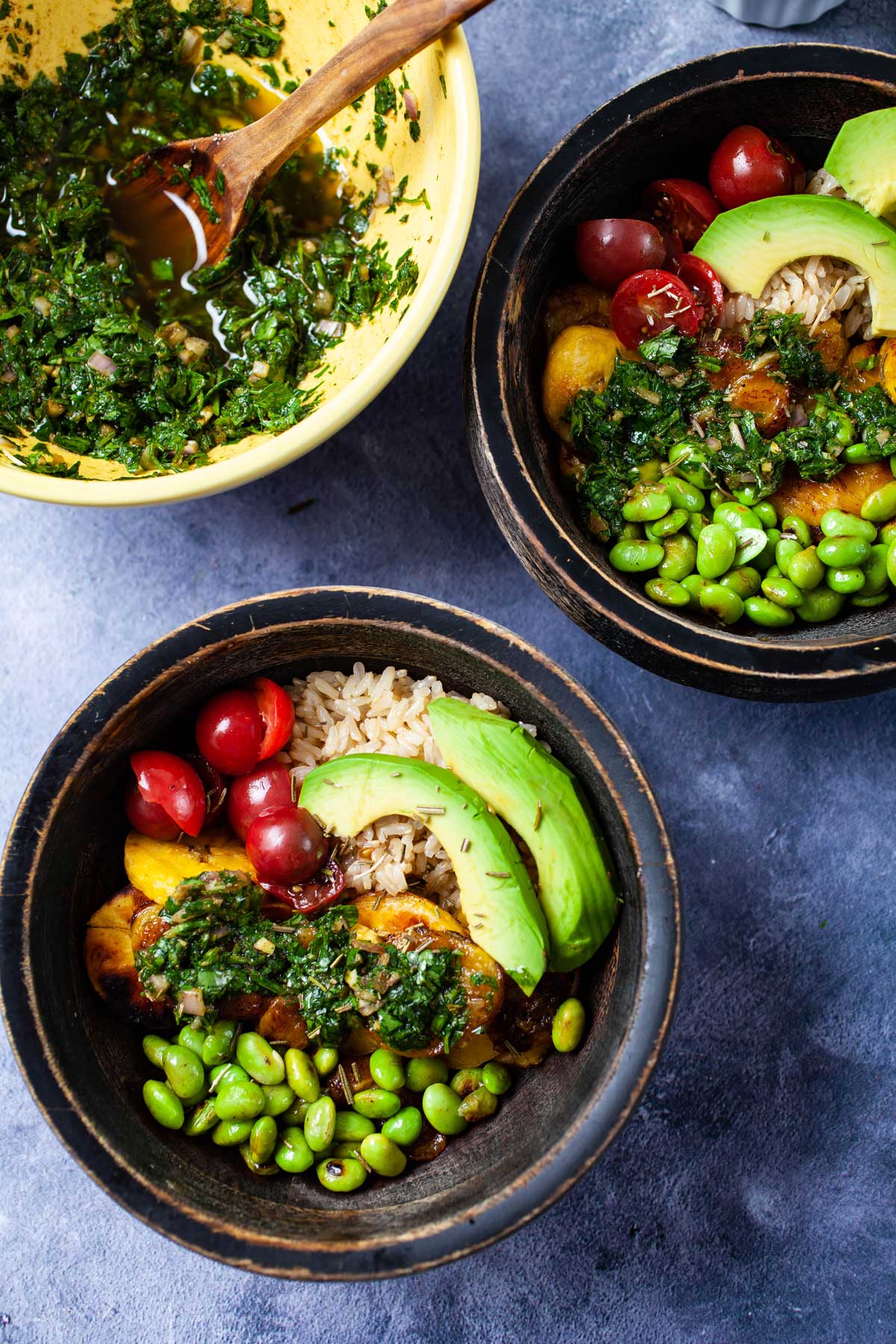 Bowls filled with cooked rice, small tomatoes, avocado slices, fried edamame beans, fried plantains, and topped with chimichurri sauce.