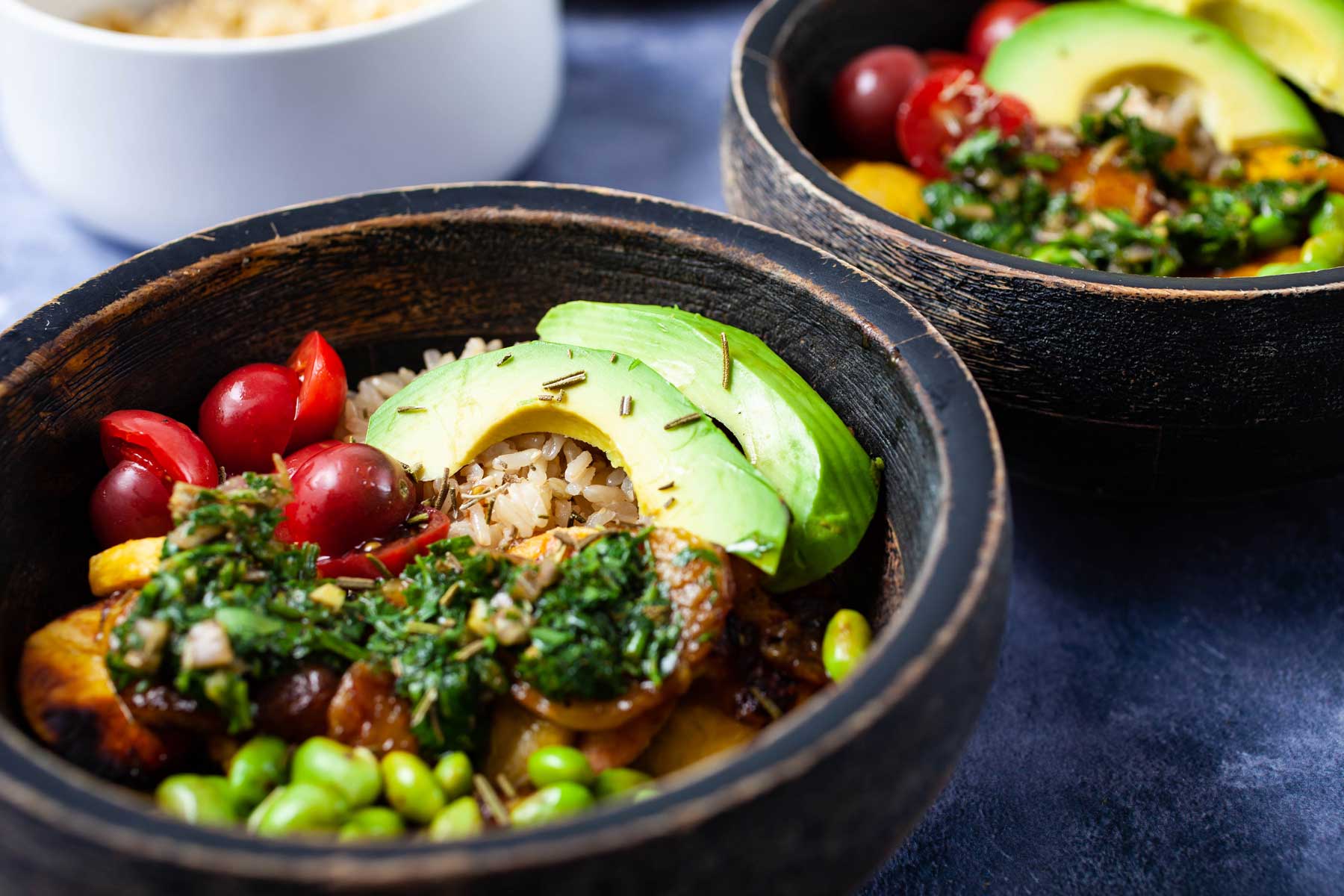 Bowls filled with cooked rice, small tomatoes, avocado slices, fried edamame beans, fried plantains, and topped with chimichurri sauce.