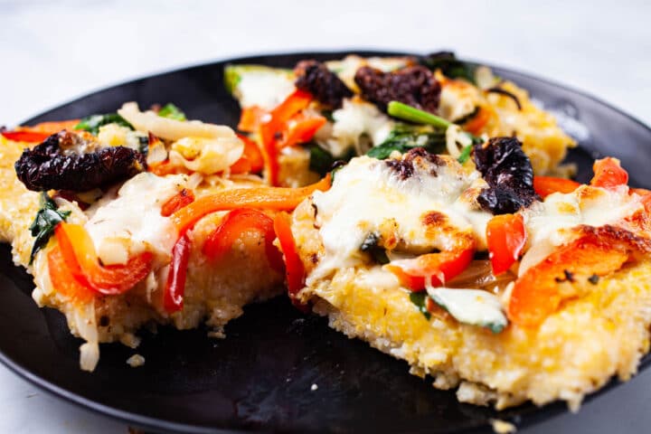 A few pieces of vegetable polenta pizza topped with spinach, red bell pepper, sun dried tomatoes, onions, garlic, and mozzarella cheese on a large plate.