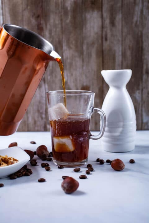 Someone pouring a cold coffee brew into a glass coffee cup filled with ice cubes and next to a milk can and nuts on the side.