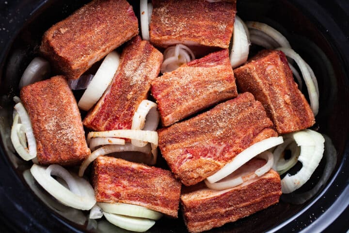 Spiced beef ribs in a slow cooker with fresh onion slices.