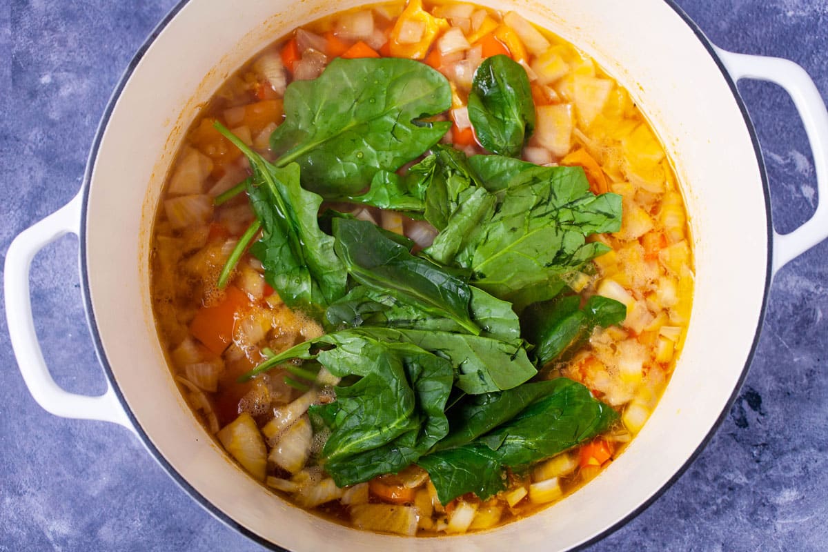 Chopped onions, bell pepper, carrots in a large pot covered with broth and topped with fresh spinach leaves.