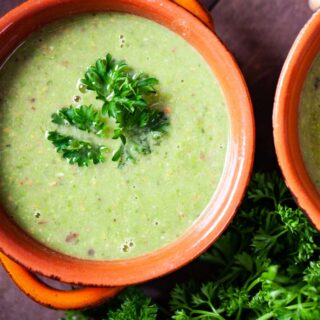 A close-up of a terra cotta bowl filled with green pea soup, topped with fresh Parsley leaves.