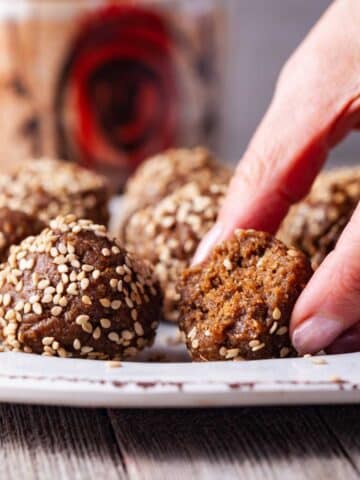 A hand placing half of an energy ball sprinkled with seeds on a small plate next to other sesame seeds sprinkled energy bites.