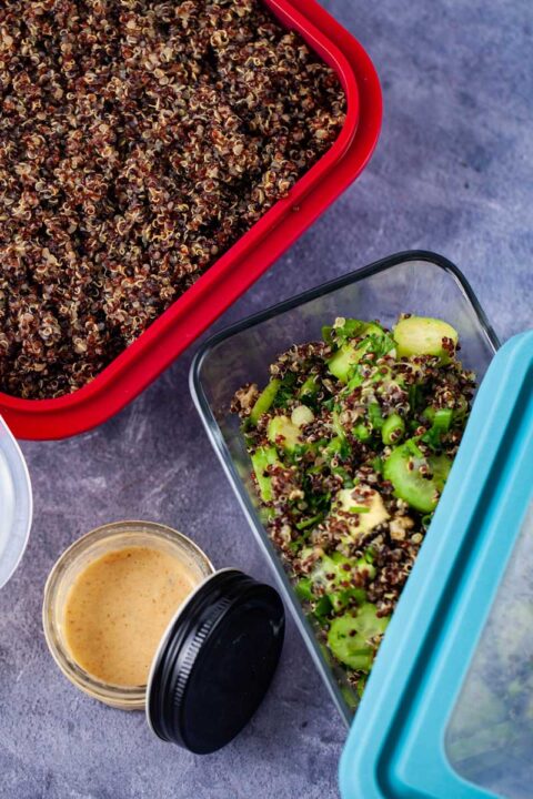 Tupperware filled with cooked red quinoa, next to a jar filled with a dressing and another container filled with avocado quinoa salad.