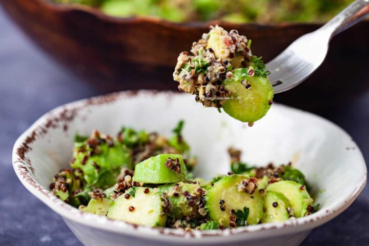 A close-up of a fork holding a big bite of cucumber quinoa salad and being lifted over a small salad bowl.