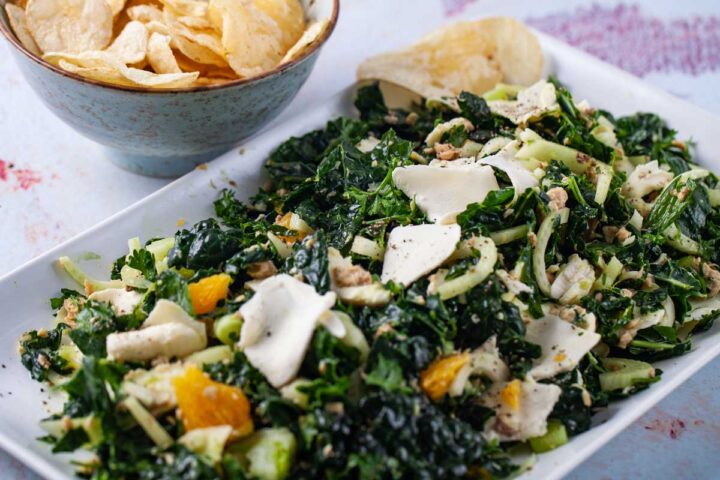 A large serving plate topped with a raw green kale and salmon salad with fennel, oranges, and mozzarella cheese, and a bowl of chips on the side.