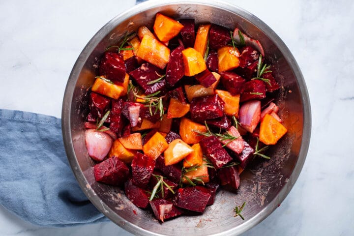 A large bowl filled with raw cubed beets, onions, and potatoes and seasoned with herbs.