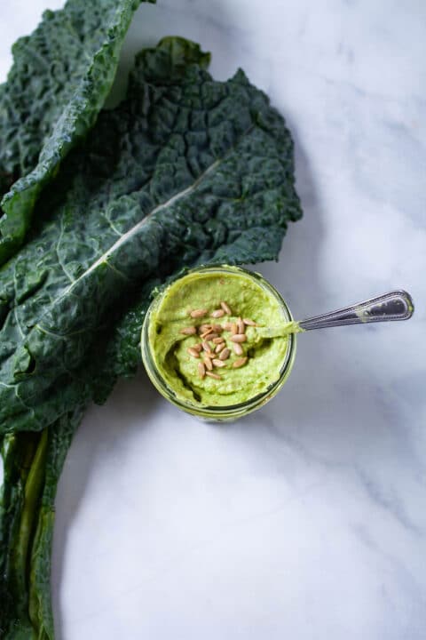 A small spoon resting in a jar filled with a green paste and topped with sunflower seeds.