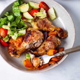 A large plate filled with three roasted chicken drumsticks, sweet potatoes, and a green salad with romaine lettuce, cucumbers, and cherry tomatoes.